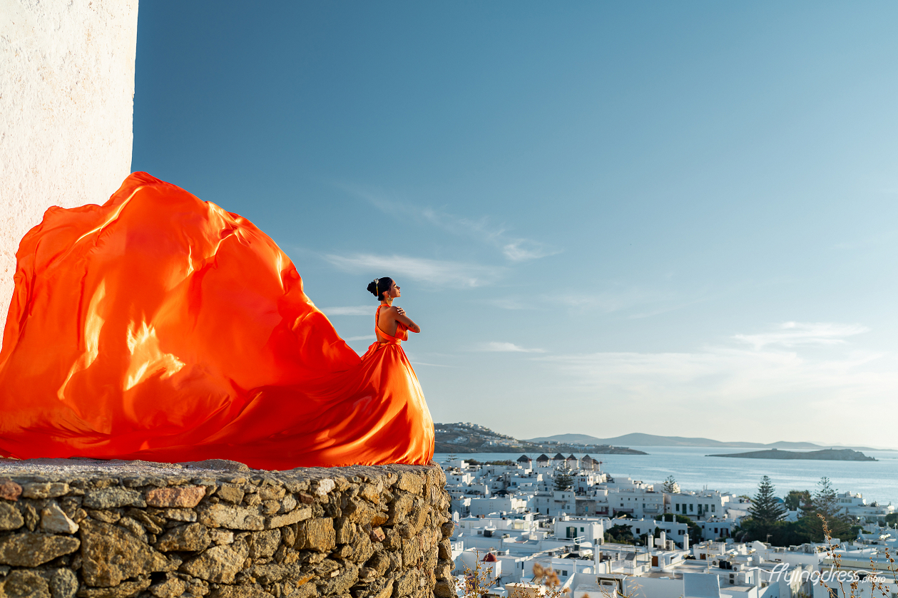 A woman in a flowing orange gown stands gracefully on a stone wall, her dress billowing in the wind against the stunning backdrop of Mykonos' white-washed buildings and the serene Aegean Sea.